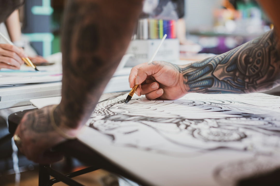 A tattooed person painting a detailed black and white design on paper